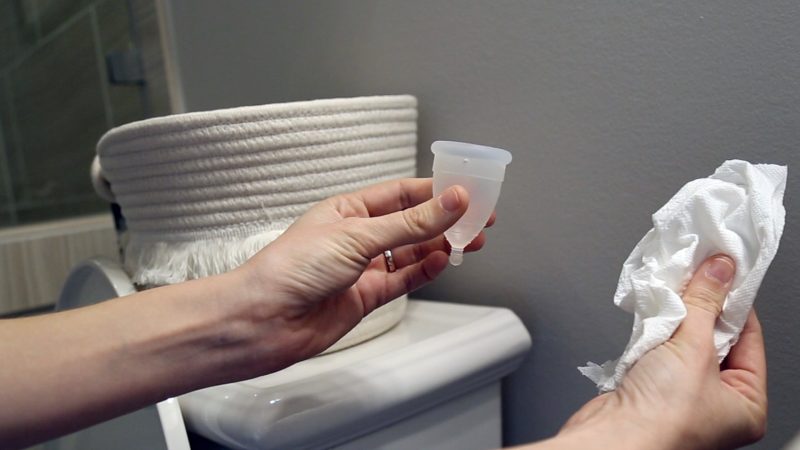 Image in a bathroom of hands holding a menstrual cup in the left hand and a tissue in the right hand