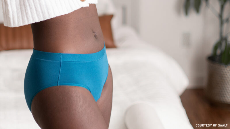Woman wearing Saalt period underwear in a vibrant blue. Lifestyle photo. A bed and plant can be seen in the background.