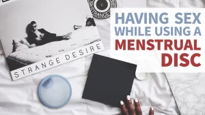 Having Sex While Using A Menstrual Disc | Image is of a bed from above. Seen in frame is a hand touching a square piece of black paper next to a menstrual disc, and an album cover that reads "Strange Desire"